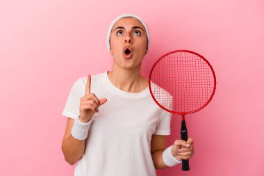 Genç, sarışın, beyaz bir kadın elinde bir badminton raketi tutuyor. Pembe arka planda izole edilmiş, ağzı açık, üst tarafı işaret ediyor..