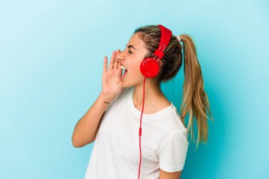 Kulaklıkla müzik dinleyen genç sarışın kadın mavi arka planda izole edilmiş bağırıyor ve avuç içini açık tutuyor..