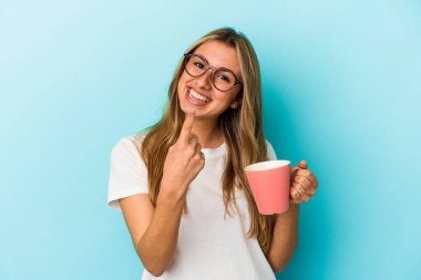 Kafkasyalı genç bir sarışın kadın mavi arka planda izole edilmiş bir kupa tutuyor ve seni işaret ediyor sanki davet ediyormuş gibi..