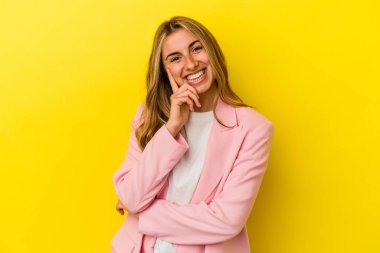 Sarı arka planda izole edilmiş genç sarışın kadın mutlu ve kendinden emin gülümsüyor, elleriyle çenesine dokunuyor..