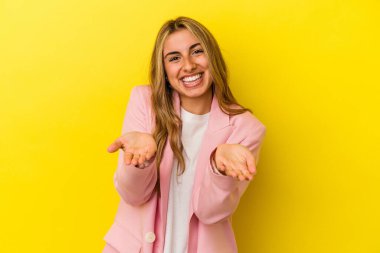 Sarı arka planda izole edilmiş genç sarışın kadın kollarıyla ölçeği yapıyor, mutlu ve kendinden emin hissediyor..