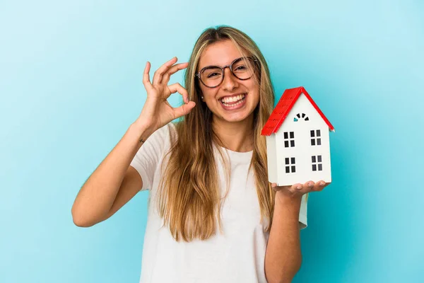 年轻的高加索女人 拿着一个蓝色背景的房子模型 快乐而自信地做出了一个好的姿态 — 图库照片