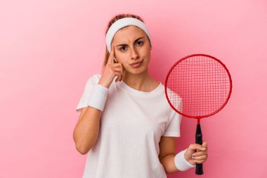 Genç, sarışın, beyaz bir kadın elinde bir badminton raketi tutuyor. Pembe arka planda izole edilmiş. Şakağı parmağıyla işaret ediyor..
