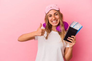 Elinde pasaport ve seyahat biletleri olan beyaz saçlı genç bir kadın pembe arka planda izole edilmiş gülümsüyor ve başparmağını kaldırıyor.