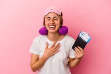 Elinde pasaport ve seyahat biletleri olan beyaz bir kadın pembe arka planda tek başına gülüyor ve elini göğsüne koyuyor..