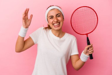 Genç, sarışın, beyaz bir kadın elinde bir badminton raketi tutuyor. Pembe arka planda izole edilmiş. Hoş bir sürpriz, heyecanlı ve el kaldırıyor..