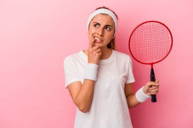 Genç, sarışın, beyaz bir kadın elinde bir badminton raketi tutuyor. Pembe arka planda izole edilmiş. Bir şeyin fotokopi odasına baktığını düşünüyor..