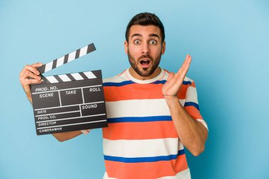 Kafkasyalı genç bir adam mavi arka planda izole edilmiş bir alçıpan tutuyor. Şaşırmış ve şok olmuş..