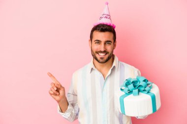 Genç beyaz adam doğum gününü pembe arka planda izole bir şekilde kutluyor gülümsüyor ve boş alanda bir şeyler gösteriyor..