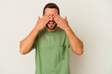 Beyaz arka planda soyutlanmış beyaz tenli genç bir adam elleriyle gözlerini kapatıyor, geniş bir gülümsemeyle bir sürpriz bekliyor..