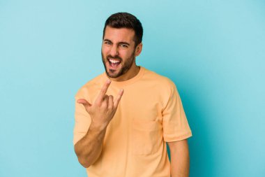 Mavi arka planda izole edilmiş genç beyaz adam parmaklarıyla kaya hareketi gösteriyor.