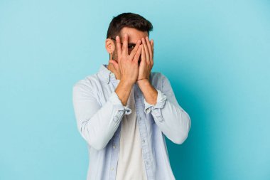 Mavi arka planda izole edilmiş genç beyaz adam kameraya parmaklarıyla göz kırpıyor, yüzünü kapatmaktan utanıyor..