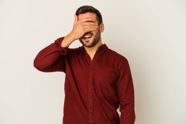 Beyaz arka planda soyutlanmış beyaz tenli genç bir adam elleriyle gözlerini kapatıyor, geniş bir gülümsemeyle bir sürpriz bekliyor..