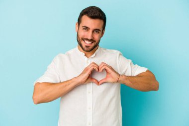 Mavi arka planda izole edilmiş genç beyaz adam gülümsüyor ve elleriyle kalp şeklini gösteriyor..