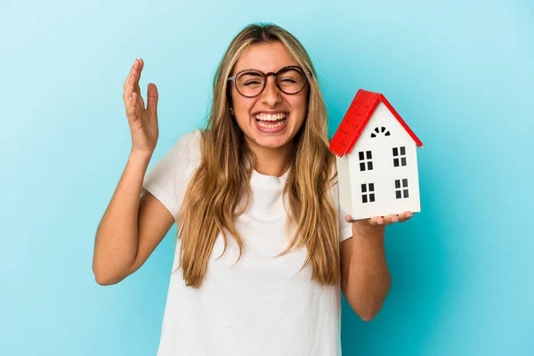 年轻的高加索女人抱着一个蓝色背景的房子模型 得到了一个愉快的惊喜 兴奋地举起双手 — 图库照片