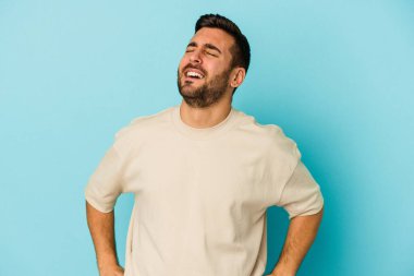 Kafkasyalı genç adam mavi arka planda izole edilmiş rahat ve mutlu bir şekilde gülüyor, boynunda dişlerini gösteriyor..