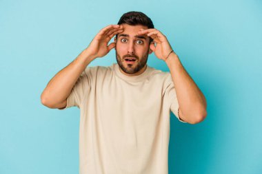 Mavi arka planda izole edilmiş genç beyaz adam hoş bir sürpriz alıyor, heyecanlı ve el kaldırıyor..