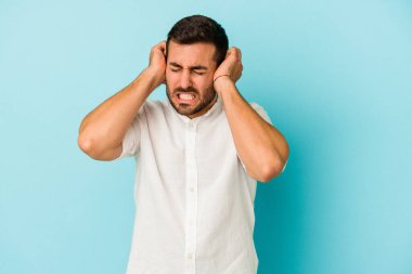Mavi arka planda izole edilmiş genç beyaz adam kulaklarını elleriyle kapatıyor..