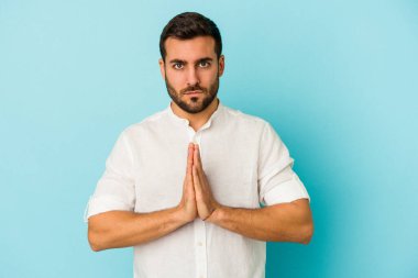 Kafkasyalı genç adam mavi arka planda izole edilmiş dua ediyor, bağlılık gösteriyor, dindar biri ilahi ilham arıyor..