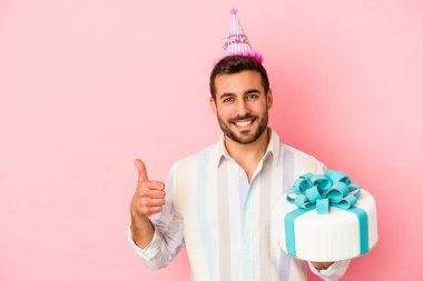 Genç beyaz adam doğum gününü pembe arka planda yalnız kutluyor gülümsüyor ve başparmağını kaldırıyor.