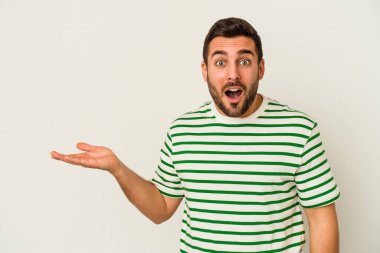 Beyaz arka planda izole edilmiş genç beyaz adam, avuç içi fotokopi alanını elinde tutmaktan etkilenmiş..
