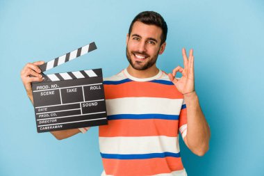 Kafkasyalı genç bir adam mavi arka planda izole edilmiş, neşeli ve kendinden emin bir şekilde el kol hareketi yapıyor..