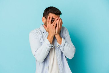 Kafkasyalı genç adam mavi arka planda izole edilmiş. Korkmuş ve gergin parmaklarıyla göz kırp..