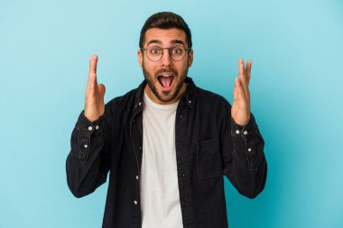Mavi arka planda izole edilmiş genç beyaz adam hoş bir sürpriz alıyor, heyecanlı ve el kaldırıyor..