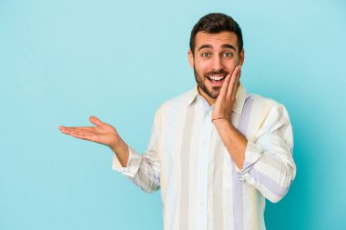 Mavi arka planda izole edilmiş genç beyaz bir adam avucunun içinde fotokopi alanı tutuyor, el üstünde tutuyor. Şaşırmış ve mutlu..