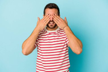 Mavi arka planda soyutlanmış genç beyaz bir adam elleriyle gözlerini kapatıyor, geniş bir gülümsemeyle bir sürpriz bekliyor..