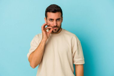 Genç beyaz adam mavi arka planda izole edilmiş ve parmakları dudaklarında bir sır saklıyor..