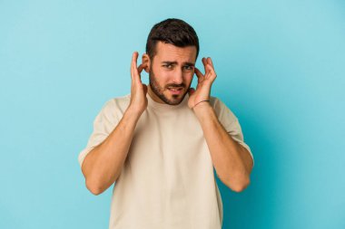 Genç beyaz adam mavi arka planda izole edilmiş parmaklarıyla kulaklarını kapatıyor, gürültülü bir ortam tarafından stresli ve umutsuz..