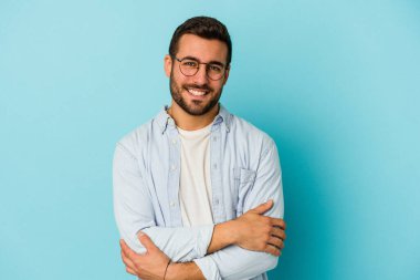 Mavi arka planda kendini güvende hisseden, kararlılıkla kollarını kavuşturan genç beyaz bir adam..