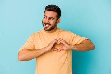 Mavi arka planda izole edilmiş genç beyaz adam gülümsüyor ve elleriyle kalp şeklini gösteriyor..