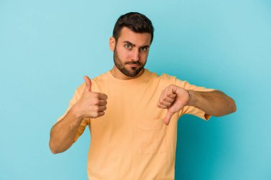Kafkasyalı genç adam mavi arka planda izole edilmiş, baş parmağını kaldırıyor, zor seçim konsepti.