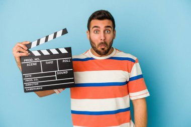 Kafkasyalı genç bir adam mavi arka planda izole edilmiş bir el çırpma tahtası tutuyor omuzlarını silkiyor ve gözleri açık..