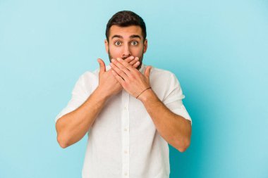 Mavi arka planda izole edilmiş genç beyaz adam elleriyle ağzını kapatırken şok oldu..