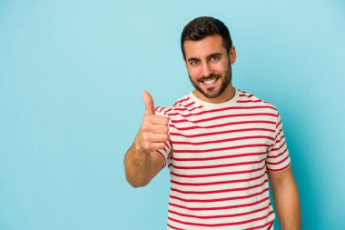 Mavi arka planda izole edilmiş genç beyaz adam gülümsüyor ve başparmağını kaldırıyor.