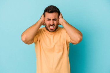 Mavi arka planda izole edilmiş genç beyaz adam kulaklarını elleriyle kapatıyor..