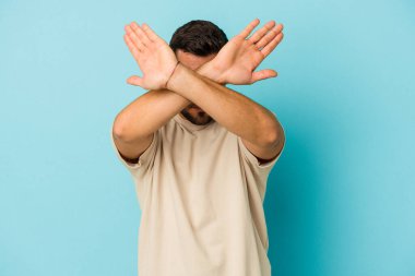 Kafkasyalı genç adam mavi arka planda izole edilmiş iki kolu çapraz tutuyor, inkar kavramı.