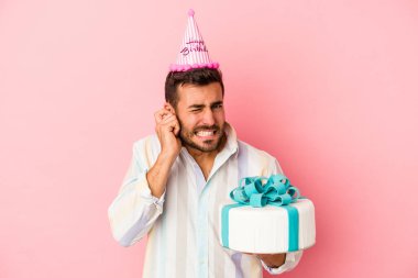Genç beyaz adam doğum gününü kutluyor. Pembe arka planda izole edilmiş. Kulaklarını elleriyle kapatıyor..