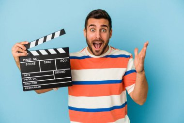 Mavi arka planda el çırpma tahtası tutan genç beyaz adam hoş bir sürprizle karşılaşıyor, heyecanlı ve el kaldırıyor..