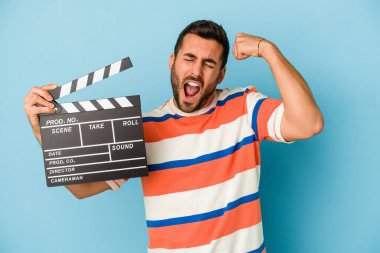 Kafkasyalı genç bir adam mavi arka planda izole edilmiş, zafer kazandıktan sonra yumruk kaldırıyor, kazanan konsept..