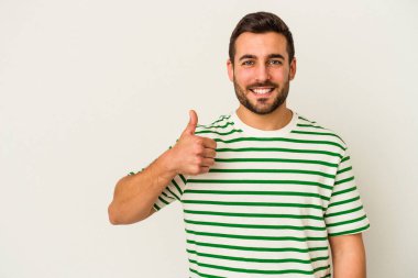 Beyaz arka planda izole edilmiş genç beyaz adam gülümsüyor ve başparmağını kaldırıyor.