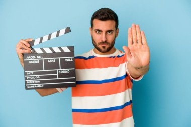 Kafkasyalı genç bir adam mavi arka planda izole edilmiş bir şekilde duruyor elini uzatmış, dur işaretini gösteriyor ve seni engelliyor..