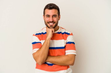 Beyaz arka planda izole olmuş genç beyaz bir adam gülümsüyor ve kendinden emin, çenesine elleriyle dokunuyor..
