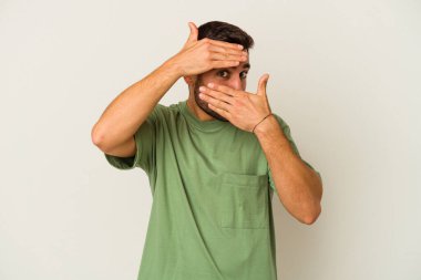 Beyaz arka planda izole edilmiş genç beyaz bir adam kameraya parmaklarıyla göz kırpıyor, yüzünü kapatmaktan utanıyor..