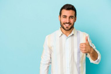 Mavi arka planda izole edilmiş genç beyaz adam gülümsüyor ve başparmağını kaldırıyor.