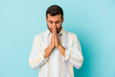 Mavi arka planda izole edilmiş genç beyaz bir adam el ele tutuşup, ağzının kenarında dua ediyor, kendinden emin..