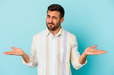 Kafkasyalı genç adam mavi arka planda izole edilmiş sorgulama jestinde şüphe ediyor ve omuz silkiyor..